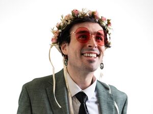 An image of the musician Will Wood wearing red tinted sunglasses, a suit, and a flower crown. He is smiling.